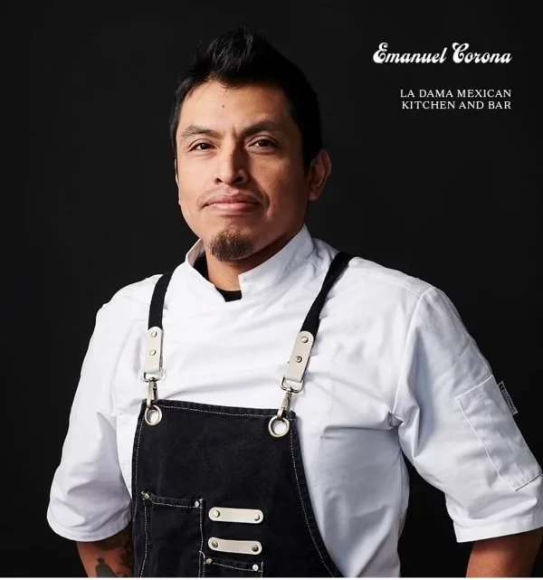 Emmanuel Corona in a white chef’s coat and dark apron against a black background.