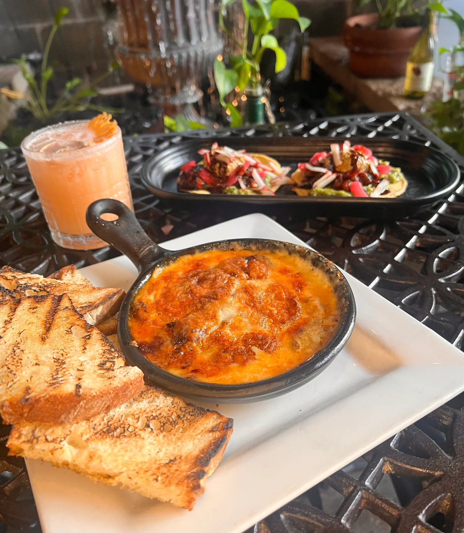 A skillet of queso fundido al pastor with grilled bread, tostadas, and a cocktail on a patio table.