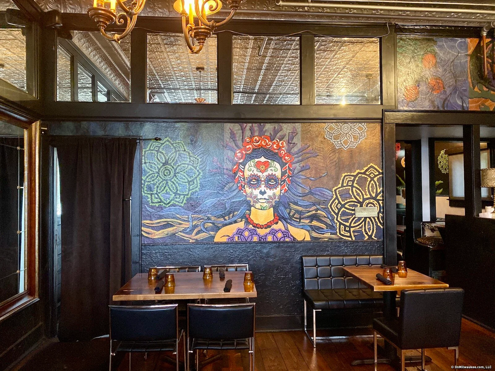 Booth tables beneath La Dama’s flower-crowned mural, with chandeliers and a patterned tin ceiling